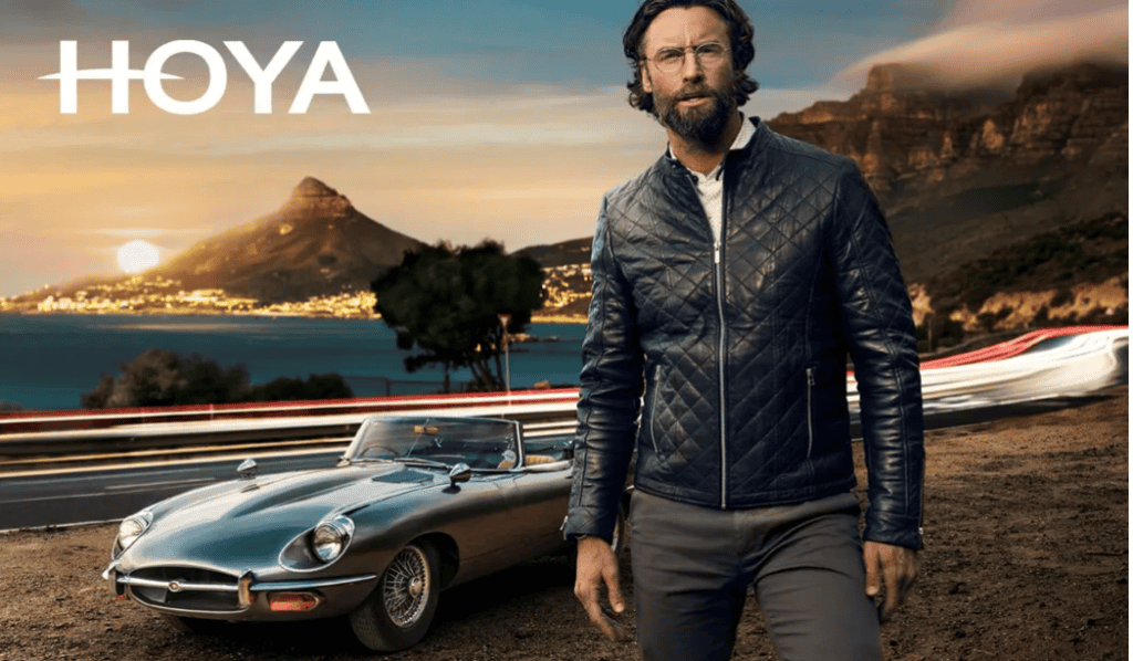 A bearded man in a quilted jacket stands in front of a silver vintage convertible sports car on a coastal road with a mountain and sunset in the background. "HOYA" is prominently displayed above.